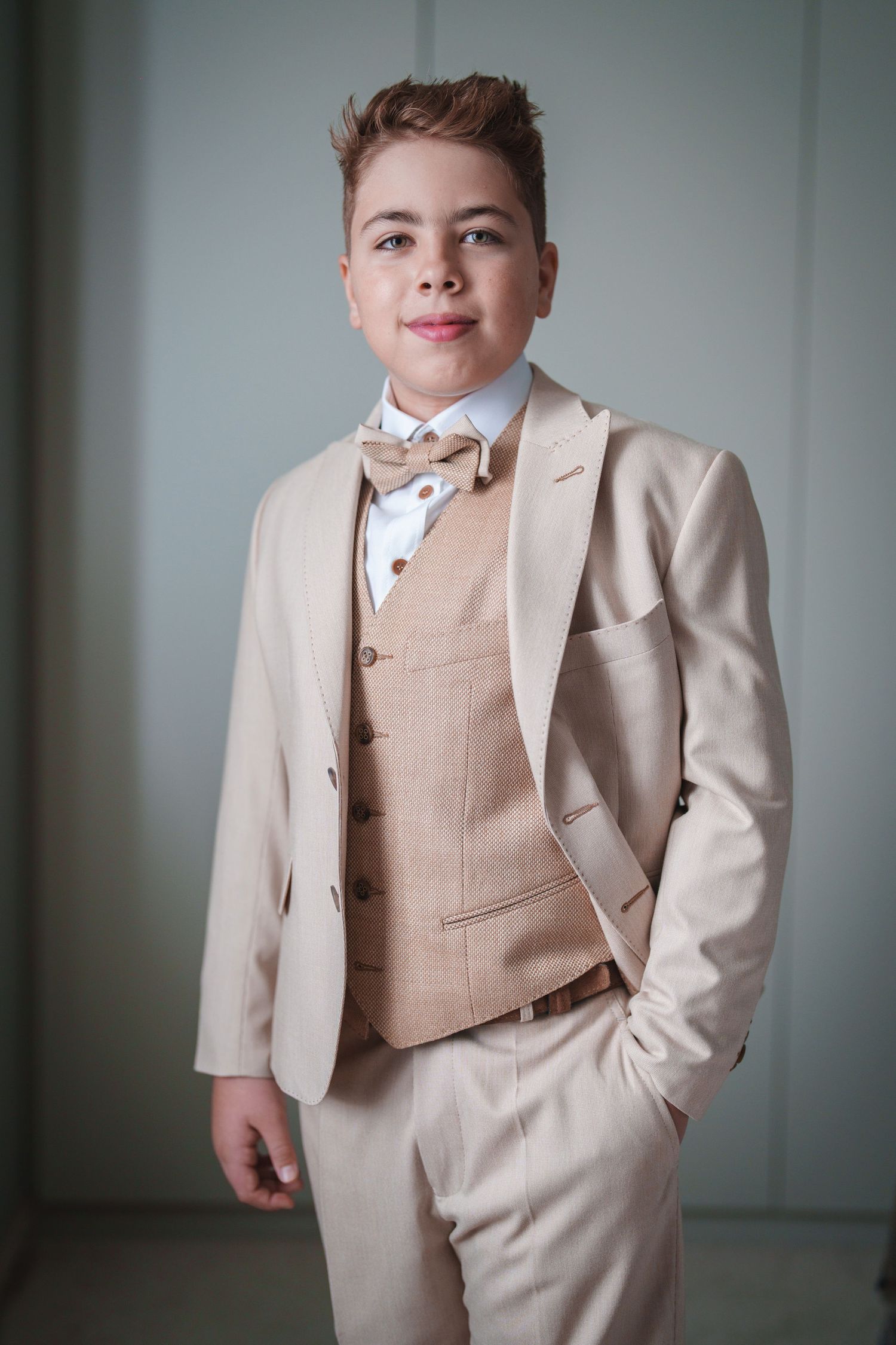 A young individual poses in a beige three-piece suit with bow tie against a light background.
