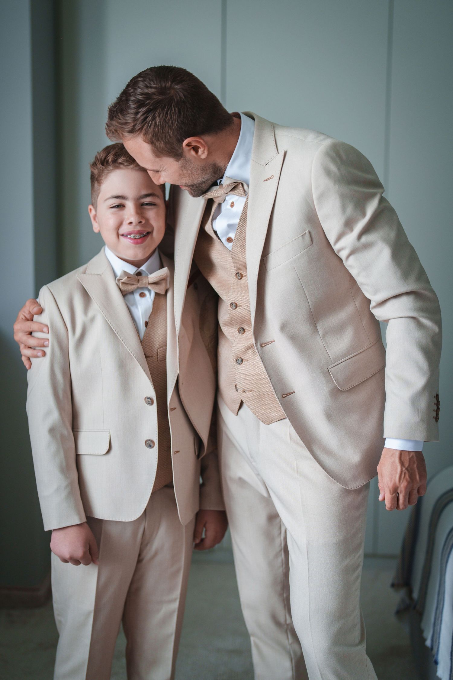 Two people in matching beige suits share a tender moment against a light wall.