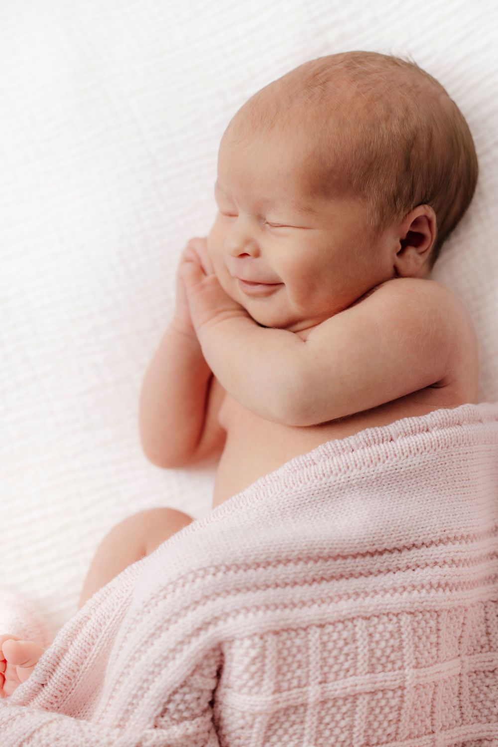 Newborn baby smiling while wrapped in pink knit blanket.