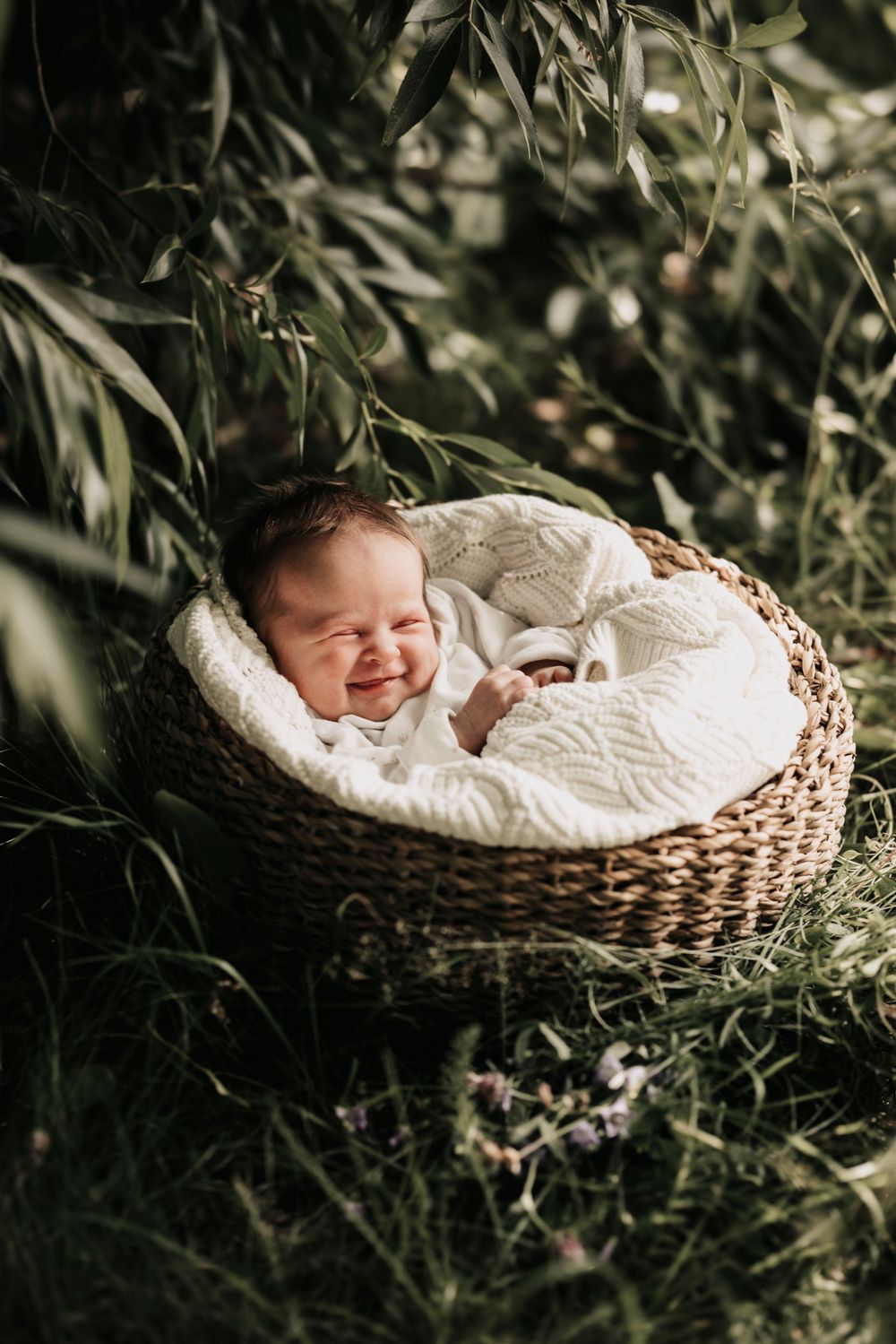 Sleeping baby in white outfit nestled in woven basket surrounded by lush greenery.