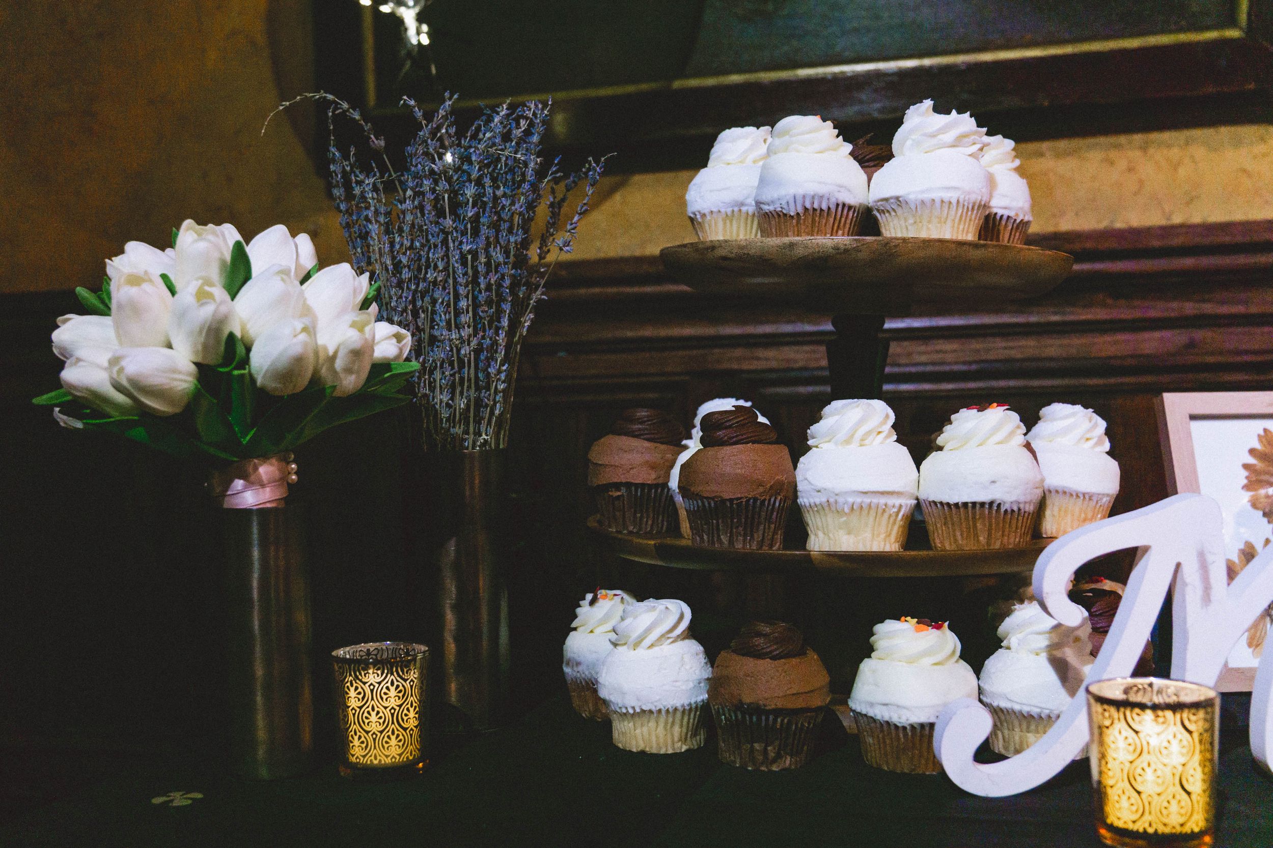 Tiered display stand with white frosted cupcakes beside white roses and golden candle holders.