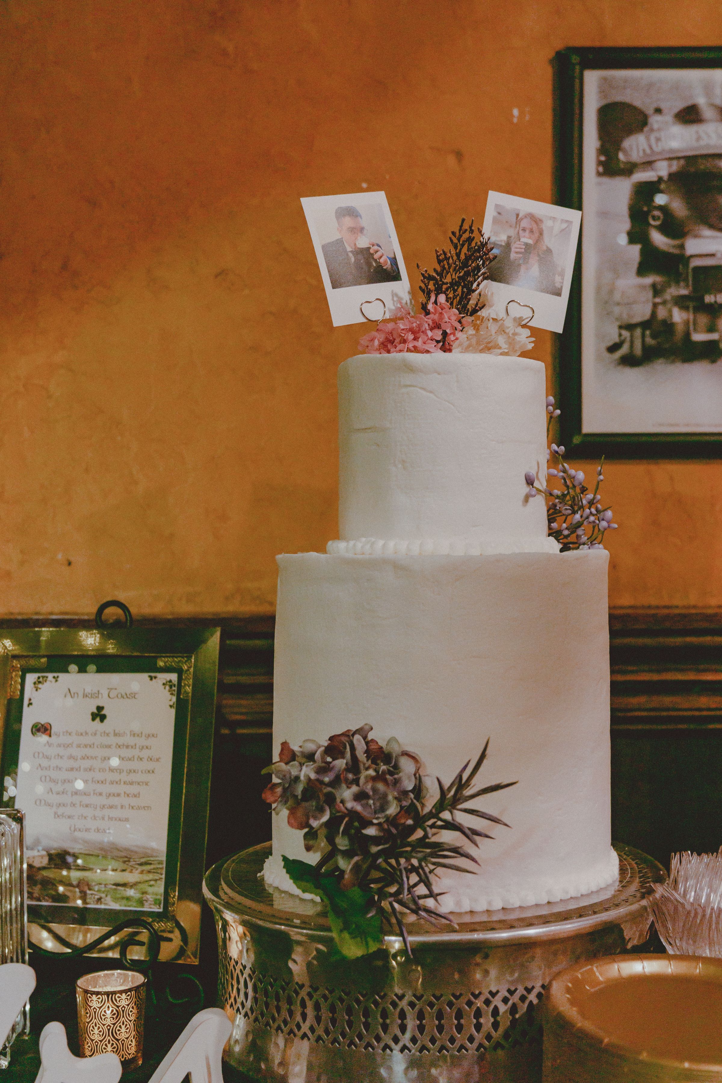 Two-tier white wedding cake topped with Polaroid photos and floral decorations on a gold stand.