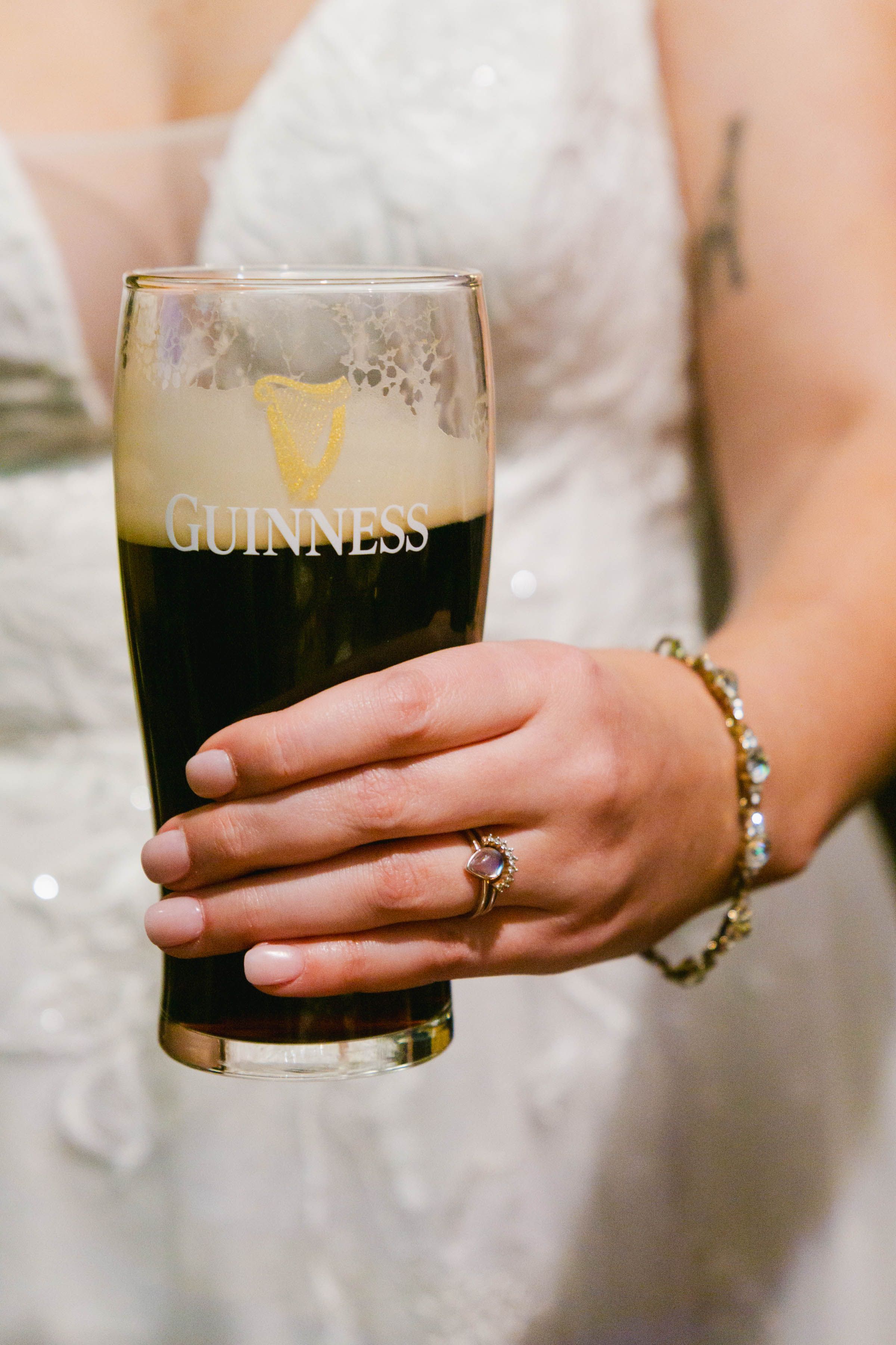 Bride in white wedding gown holding a pint of Guinness beer, showing engagement ring and bracelet.