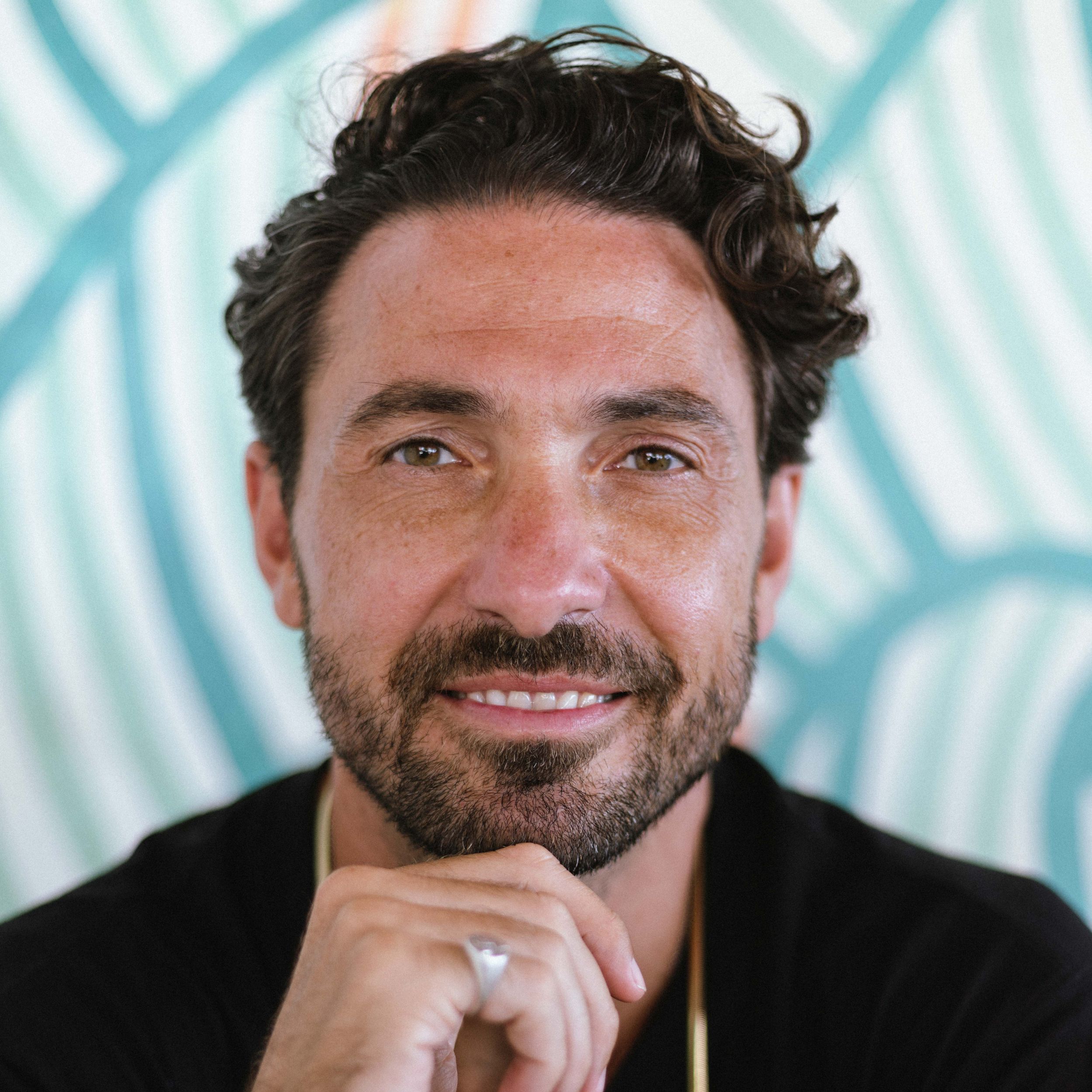 Professional headshot of person with dark curly hair against blue patterned background.