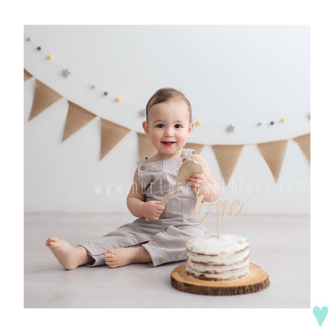 Baby in gray outfit sits on floor with white cake and burlap bunting banner decoration.