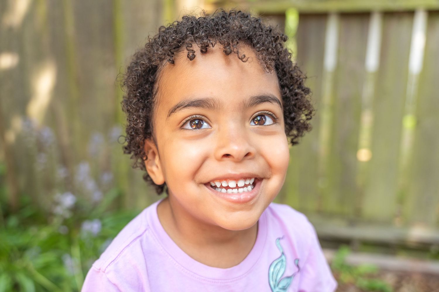 Portrait of a child in purple shirt smiling outdoors with natural background.