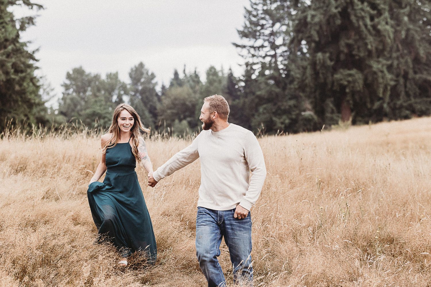 A couple holds hands while walking through a sun-lit golden field with evergreen trees in the background.