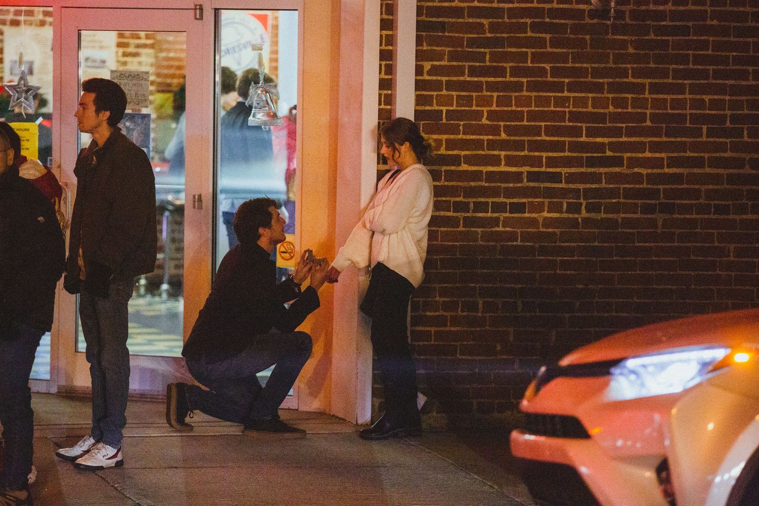 A romantic outdoor proposal scene at night in front of a brick wall and illuminated storefront.
