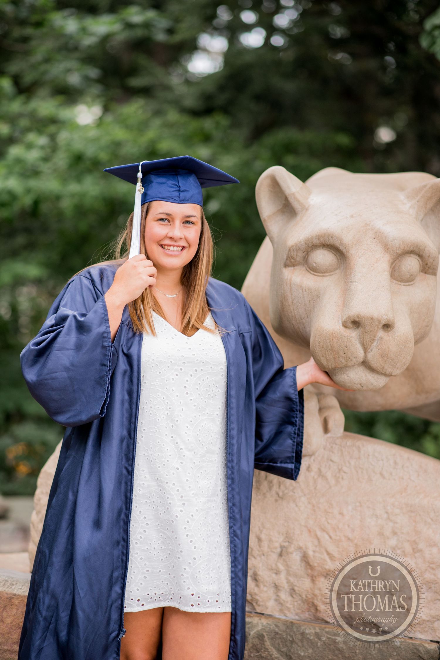 Graduate in blue cap and gown poses cheerfully beside stone lion statue at Penn State.