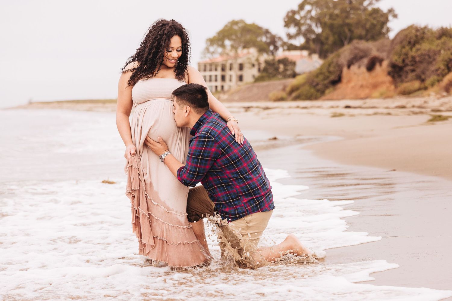 Sweet moment captured on sandy beach with gentle waves and coastal cliffs in background.