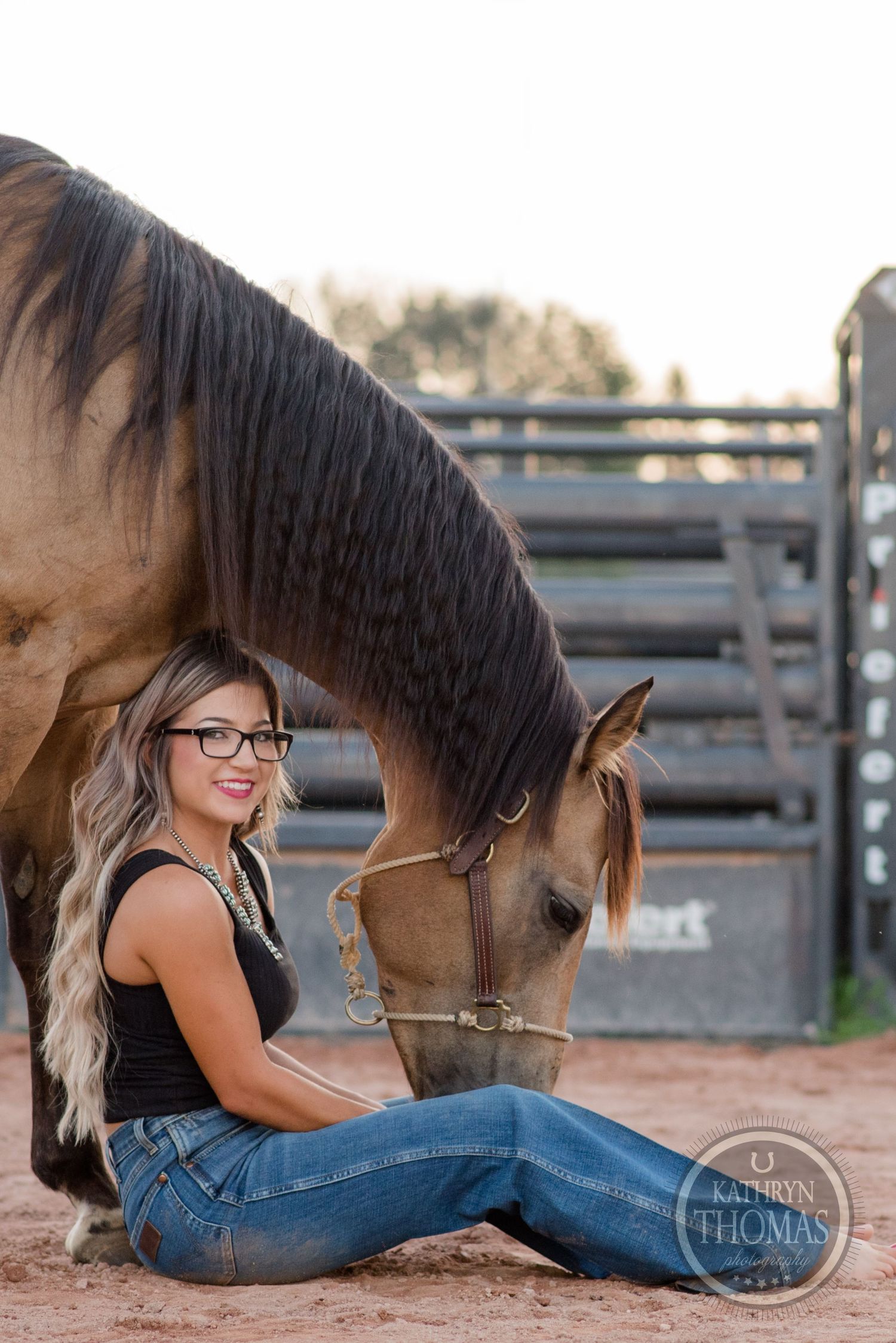 Horse lowers head to connect with handler sitting on ground by metal gates.