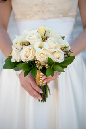 Classic white rose bridal bouquet with green foliage held against white wedding dress.