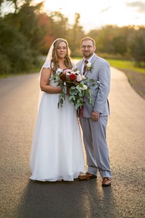 Newlyweds posing on road at sunset with bride holding burgundy bouquet.