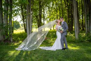 Romantic wedding portrait with flowing veil in a sunlit wooded setting.
