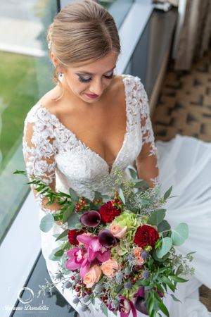 A bride in lace dress holds colorful wedding bouquet with pink and red flowers.