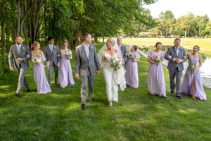 Wedding party in gray suits and lavender dresses walking outdoors on a sunny day.
