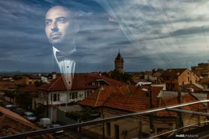 Large portrait overlaid on cityscape with orange rooftops and cloudy sky.