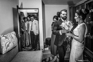 Black and white wedding photo showing groomsmen peeking through doorway as couple exchanges flowers.
