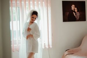 Bride in white robe and veil poses by window during wedding morning preparations.