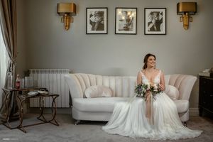 Elegant bridal portrait featuring a white wedding dress and bouquet against a gray room with wall sconces and framed artwork.