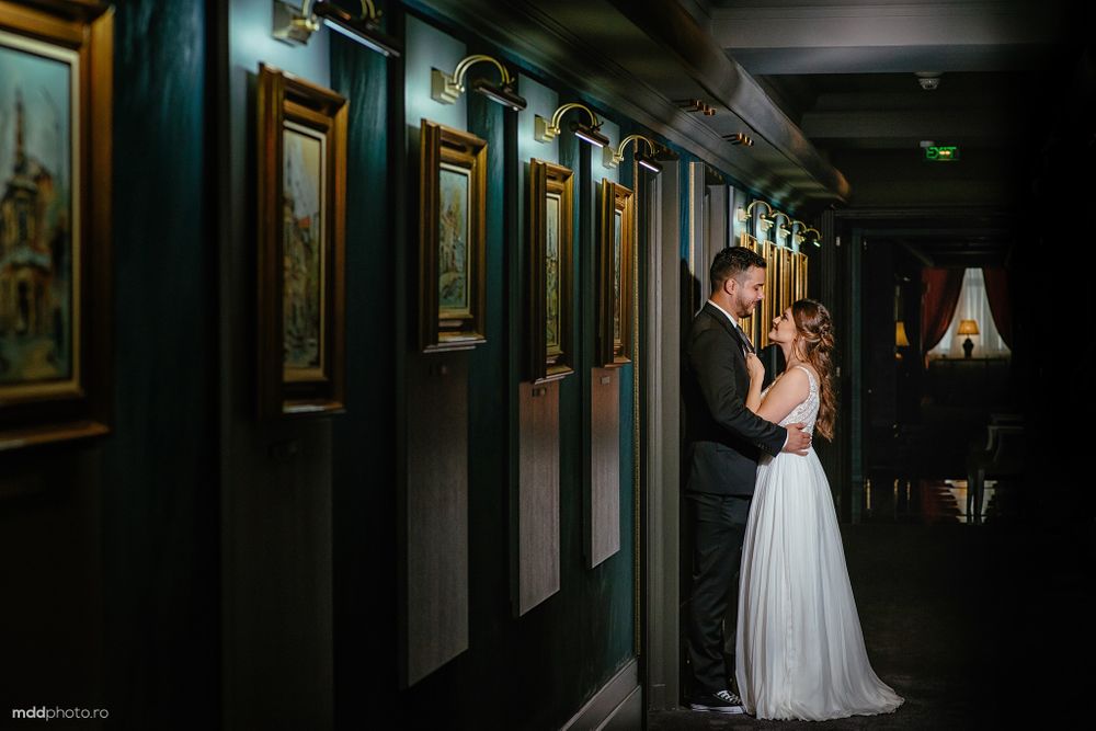 Romantic couple sharing intimate moment in dimly lit hallway with vintage artwork and moody lighting.