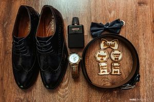 Wedding accessories including black dress shoes, bow tie, watch and perfume arranged on wooden surface.