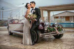 Wedding couple poses with vintage car and floral bouquet against industrial backdrop.