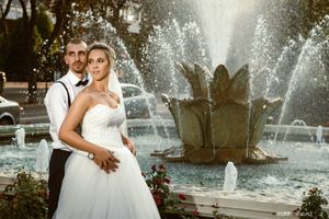 A romantic wedding portrait of a couple posing in front of an ornate fountain with water sprays in the background.