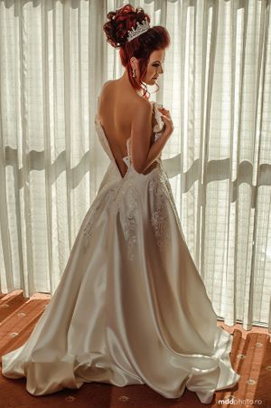 Back view of a wedding dress with intricate lace details and open back design against sheer curtains.