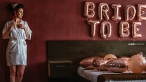 White robe and 'Bride to Be' balloon letters create a romantic pre-wedding scene against a burgundy wall.