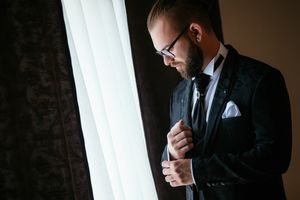 Groom in formal attire adjusts cufflinks by window in moody lighting.
