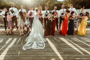 Wedding party celebrates on wooden deck during sunset with colorful attire and flowing veil.