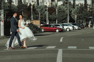 Wedding couple crosses urban street together with parked cars in background.