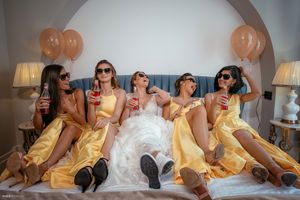 Bridal party in yellow dresses celebrates on bed with champagne and balloons.
