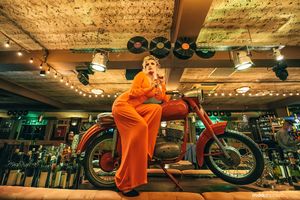Woman in orange jumpsuit posing on vintage motorcycle in dramatically lit indoor setting.