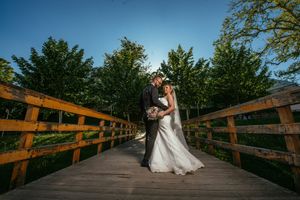 Romantic wedding photo on a wooden bridge surrounded by green trees at dusk.