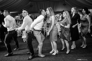 A black and white photo of wedding guests dancing in a line at a reception party.