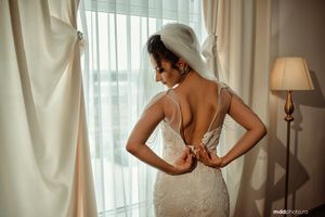 Back view of a bride adjusting her fitted lace wedding dress near a window.
