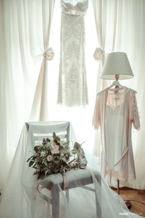 Wedding dress and robe hanging by window with bridal bouquet and lamp on side table.