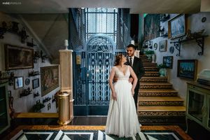 Wedding couple standing at base of ornate staircase with vintage decor and artwork.