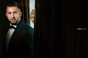 Moody portrait of groom in emerald velvet tuxedo jacket standing by window.