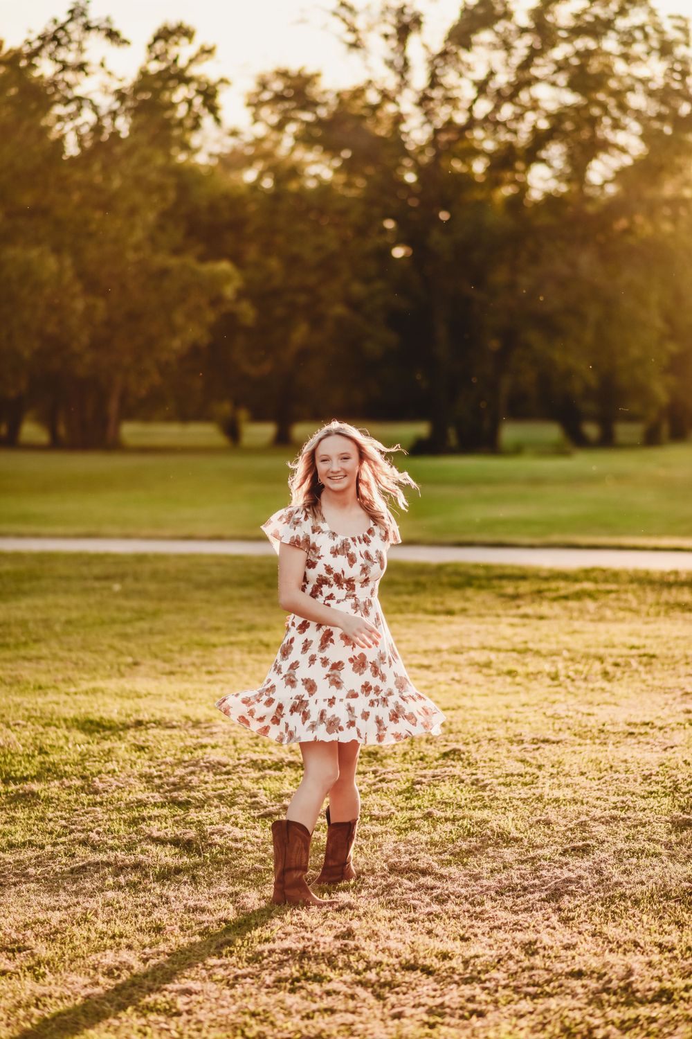 A floral dress and brown boots twirl in golden sunlight on a grassy field during sunset.
