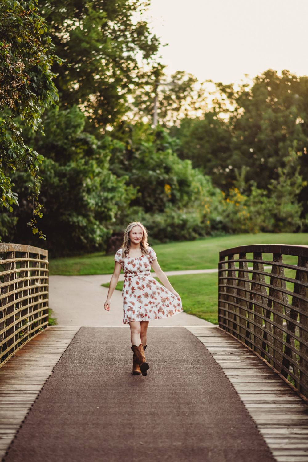 Floral dress flows while walking across wooden bridge pathway surrounded by greenery.