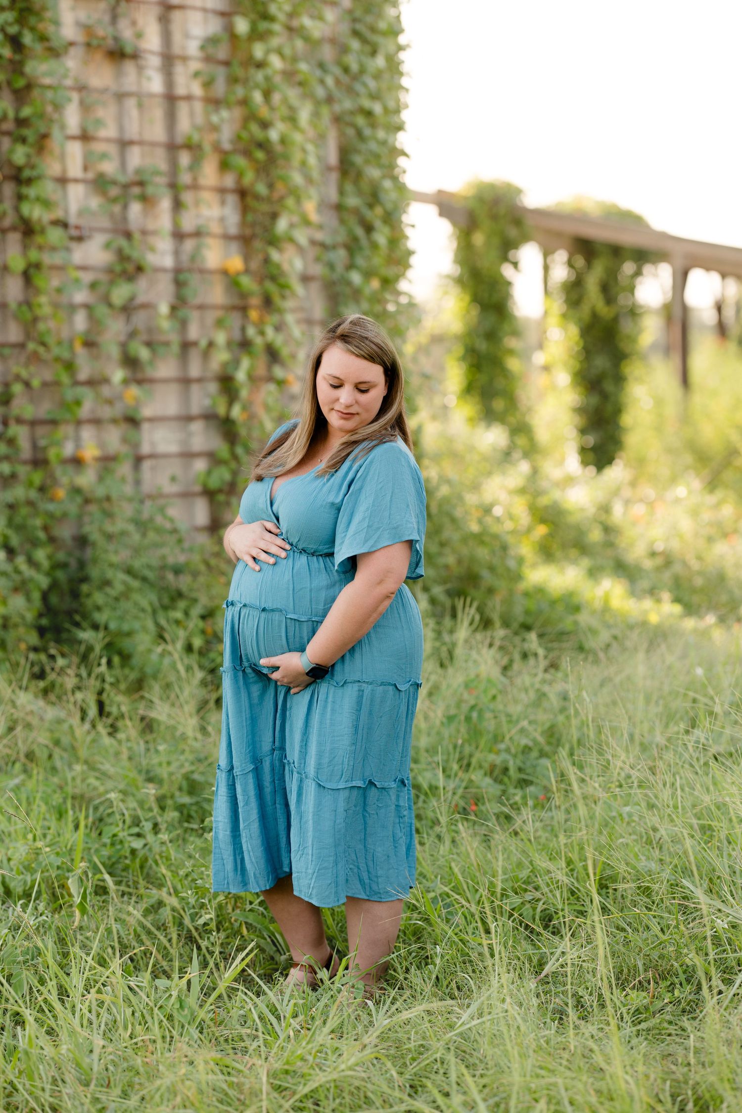 Pregnant woman in blue dress standing by ivy-covered wall in garden setting.