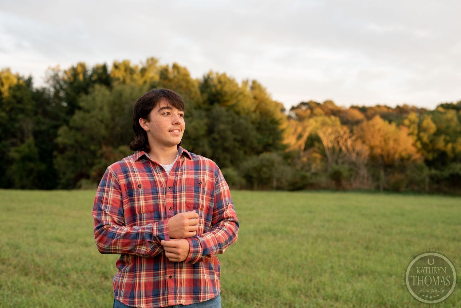 Person in plaid shirt stands in field with autumn trees and golden light in background.