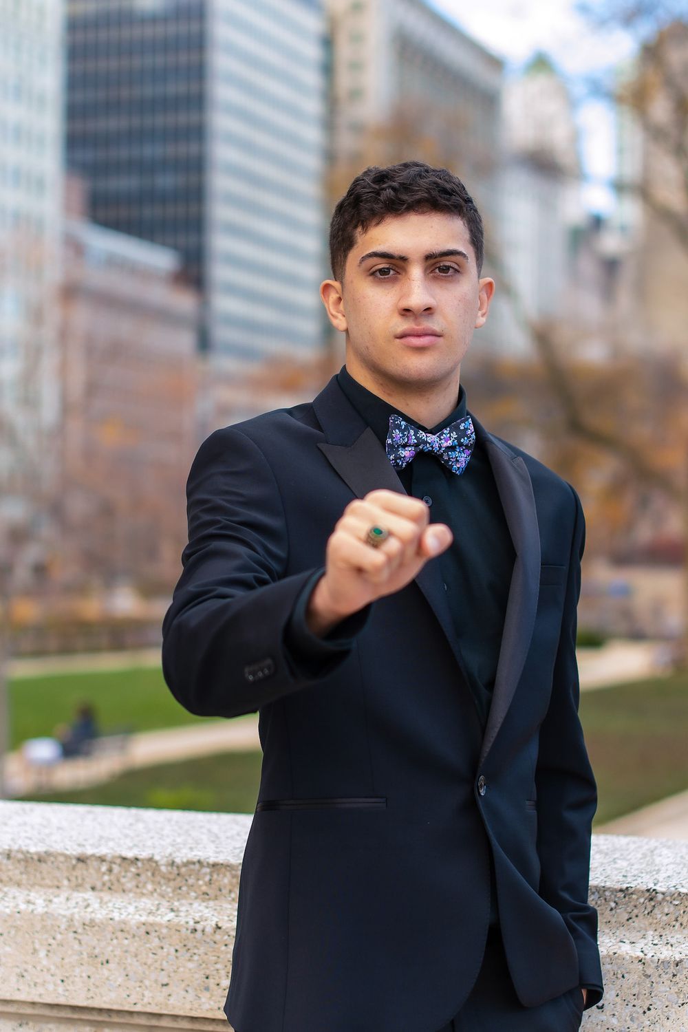 Person in black suit with bow tie making expressive hand gestures against urban background.