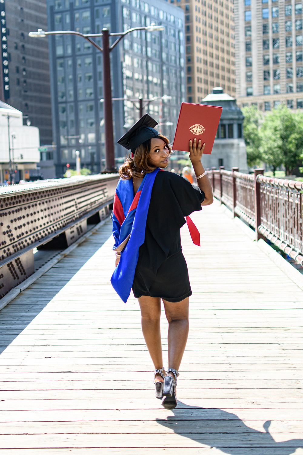 Graduate in cap and gown poses on wooden bridge with city skyline background.