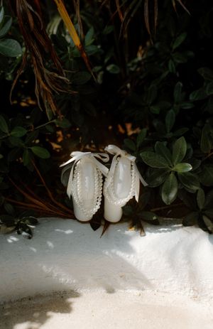 Small white baby shoes with ribbons sitting on white sand against dark foliage background.