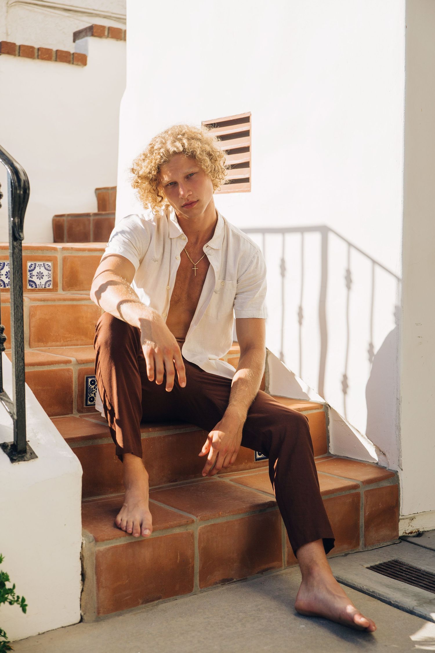 Curly blonde male model sitting on Spanish tile steps in casual white Vissla button-down shirt, San Clemente beach fashion editorial