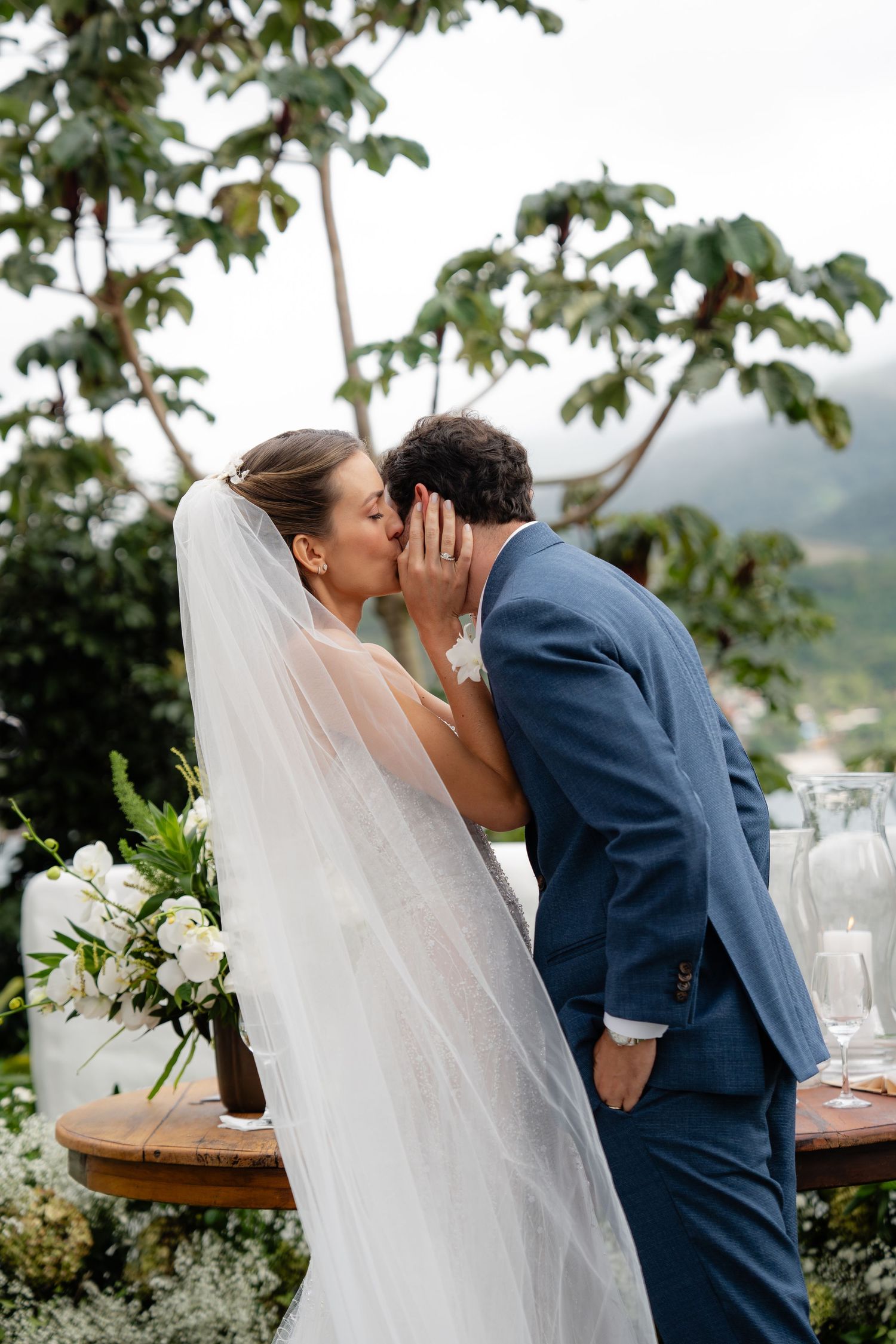 Intimate whispered moment between bride in flowing veil and groom at outdoor reception.